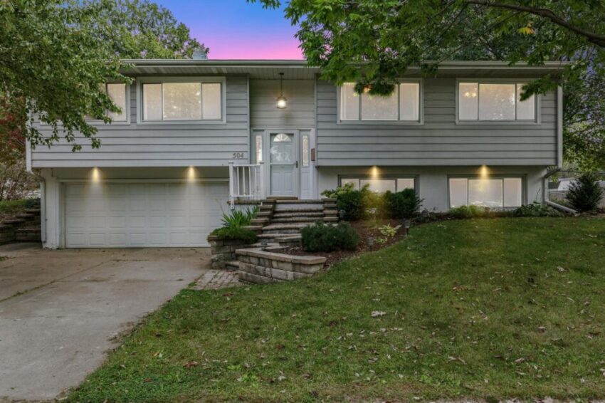 Home with glowing windows and concrete driveway with mature trees.