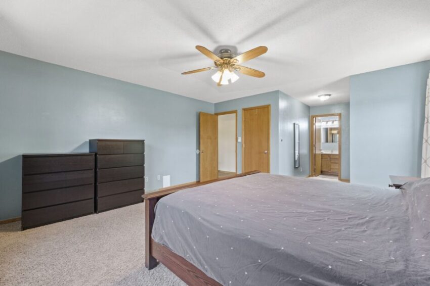 Bedroom with blue walls, ceiling fan, bed, dressers, and open door to bathroom.