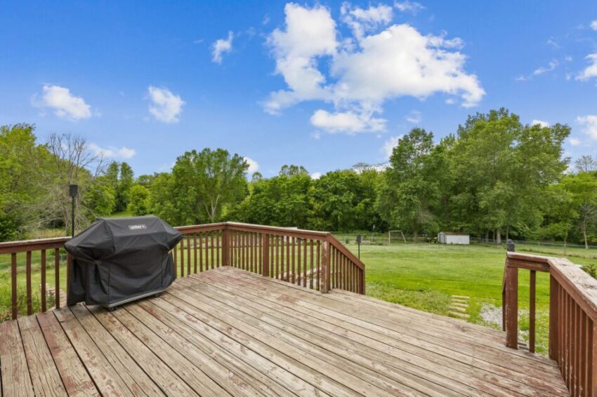Wooden deck with grill overlooking grassy yard and trees under a blue sky.