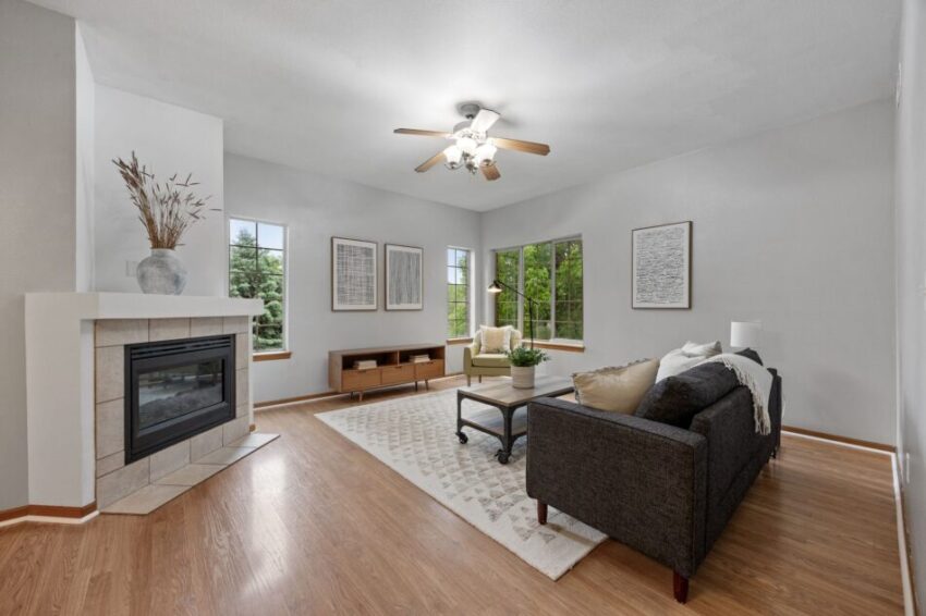 Bright living room with fireplace, gray sofa, and large windows overlooking greenery.