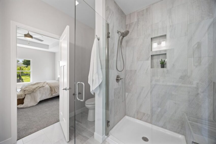 Modern bathroom with glass shower, white towel, and view into bedroom.