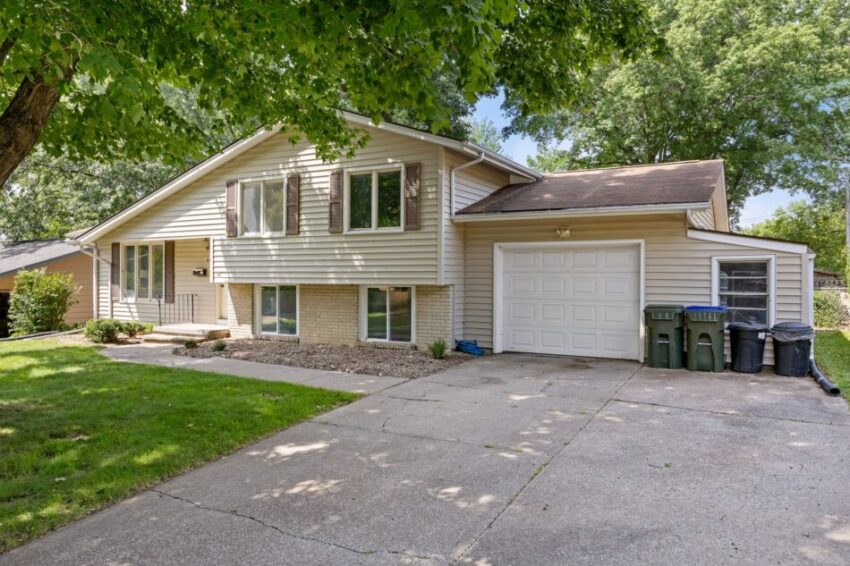 Suburban house with garage, driveway, and green lawn under large trees.
