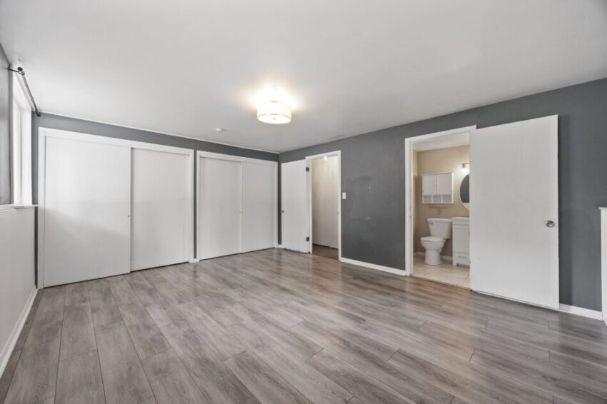 Spacious empty room with gray walls, wooden floor, and open door to bathroom.