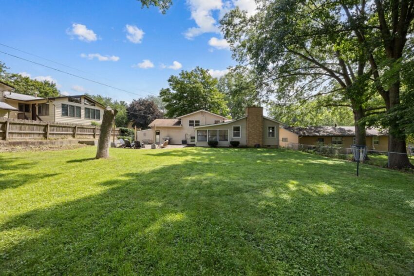 Spacious backyard with green lawn, trees, and a house under a blue sky.