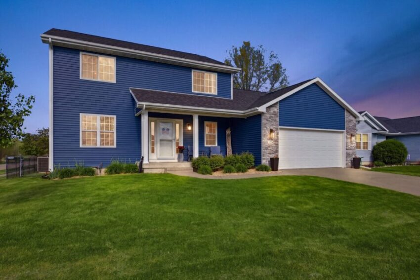 Two-story blue house with lit windows, front porch, and well-maintained lawn at dusk.
