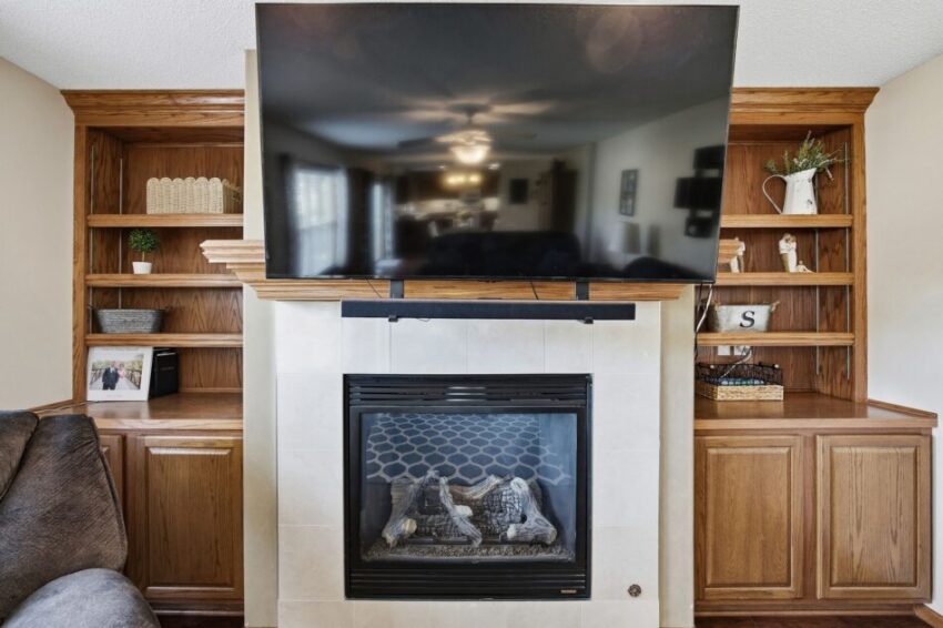 Living room with mounted TV above fireplace, wooden shelves, and cabinets on both sides.