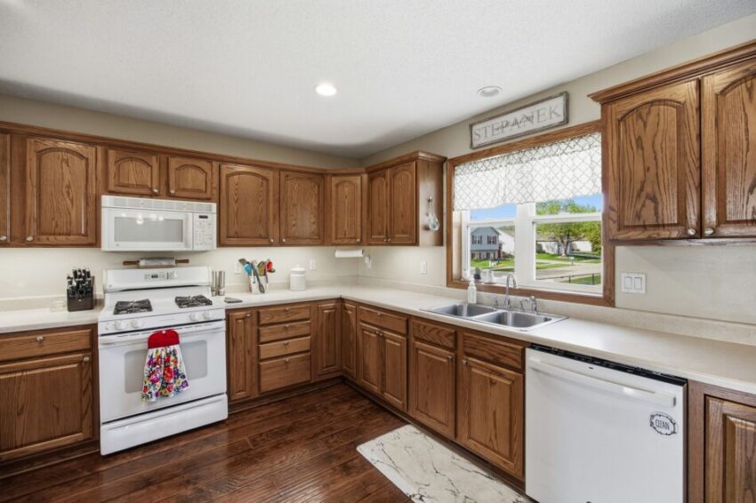 Spacious kitchen with wooden cabinets, white appliances, large window, and hardwood floors.
