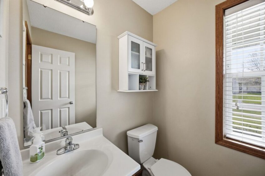 Small bathroom with sink, toilet, wall cabinet, mirror, and window with blinds.