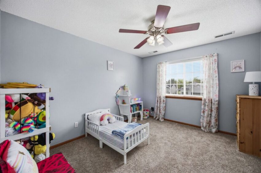 Children&rsquo;s bedroom with toddler bed, toys, bookshelf, window with curtains, and ceiling fan.
