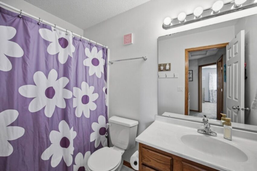 Bathroom with purple floral shower curtain, white toilet, wooden vanity, and large mirror.