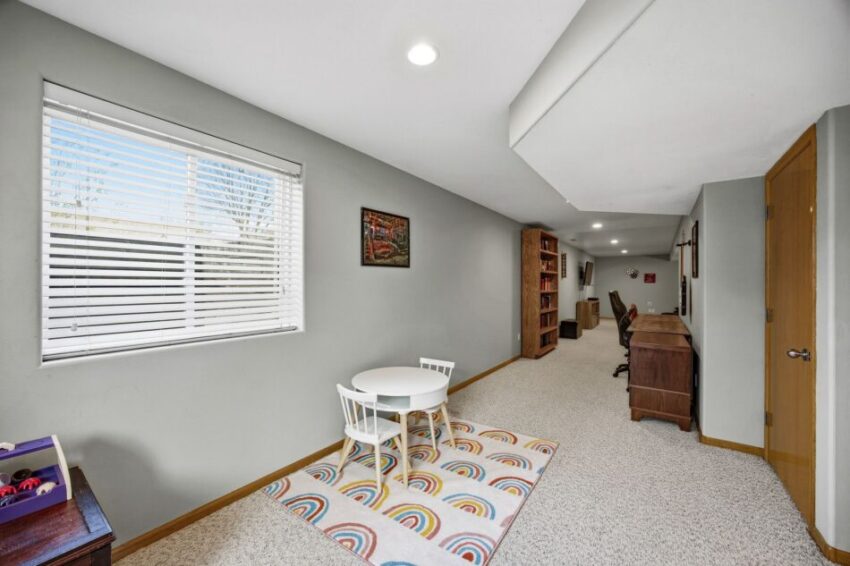 Basement room with play area, small table, bookshelves, desks, and window with blinds.