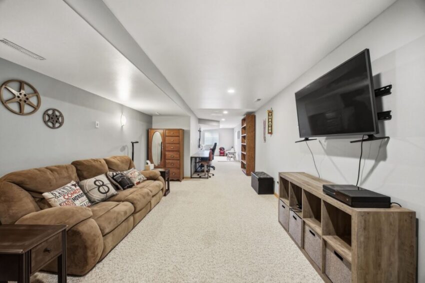 Spacious basement with brown sofa, wall-mounted TV, storage shelves, and home office area in background.