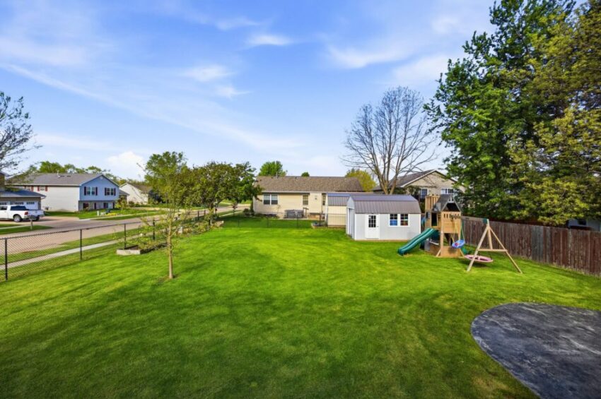 Spacious backyard with green lawn, playground set, shed, and surrounding houses under blue sky.