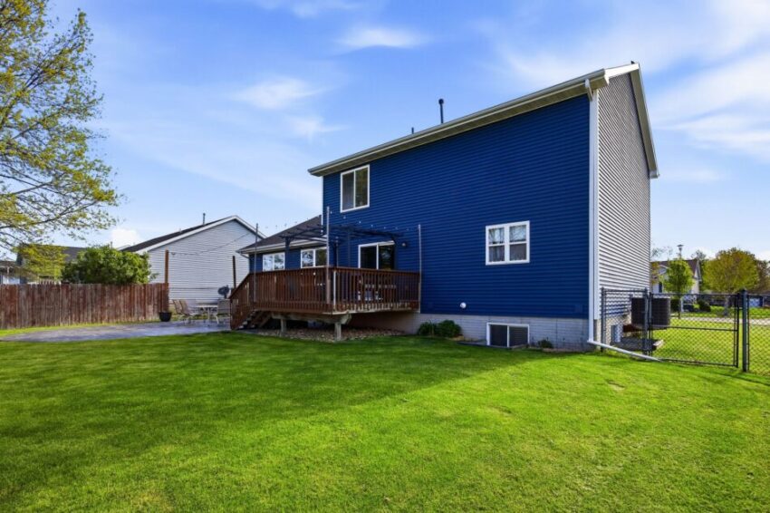Back view of blue two-story house with wooden deck and fenced green yard.